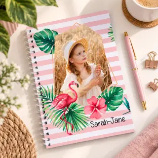 A personalised spiral-bound notebook with a custom photo cover featuring a couple, placed on a white desk next to a keyboard, a black and white pen, push pins, and a small green plant.